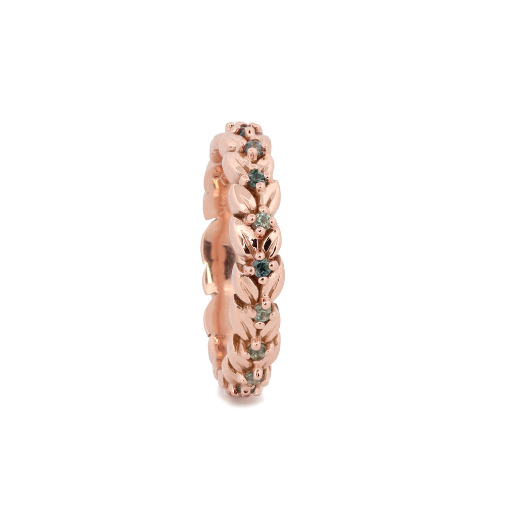 A rose gold eternity ring featuring a detailed floral pattern with small green gemstone accents, shown on a plain white background.
